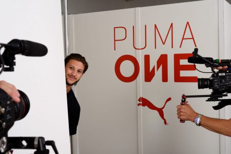 Two cameras point at a man peeking out from behind a wall with the Puma One logo on it
