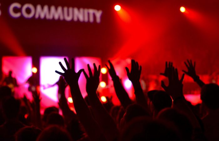 The silhouette of many hands in the air, behind them red, party lights and a sign saying "community"