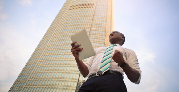 A businessman holding an iPad, clenching his fist and looking motivated, a highrise building behind him